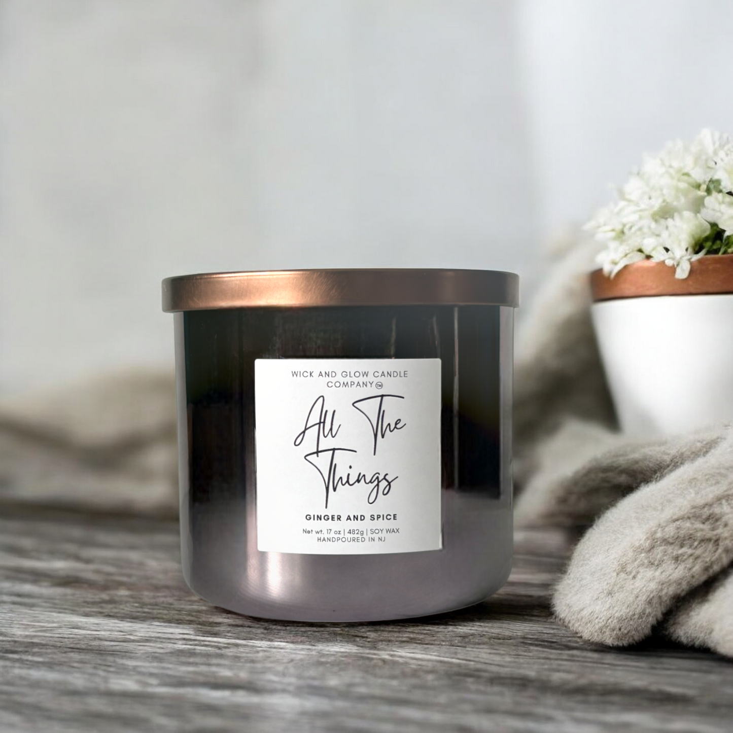 Wick and Glow luxury soy wax candle in a gray glass container with a bronze lid on a gray wooden table with a plant in the background. The scent is Ginger and spice