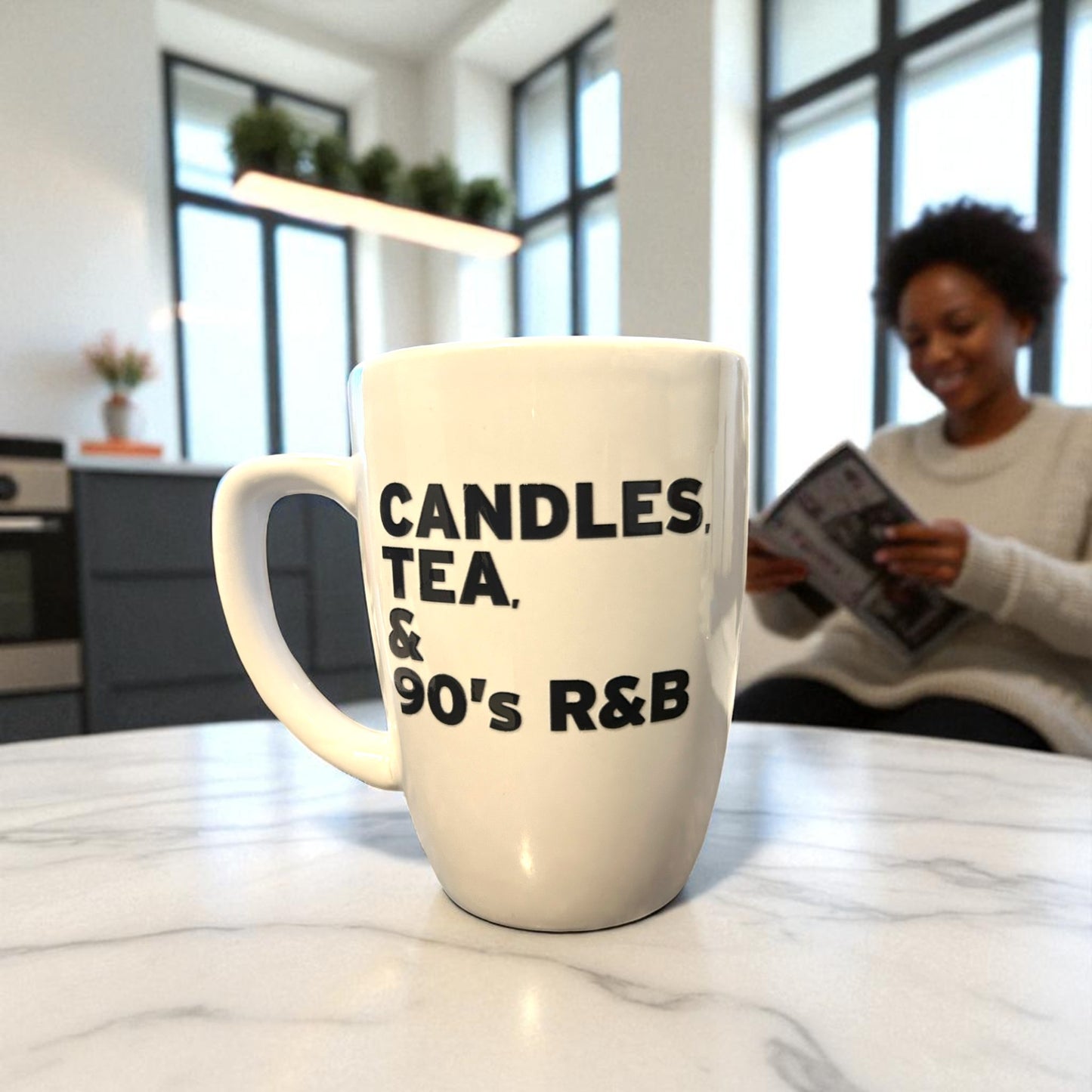 White mug with text 'CANDLES, TEA, & 90's R&B' on a marble countertop with a blurred background of a person reading a book.