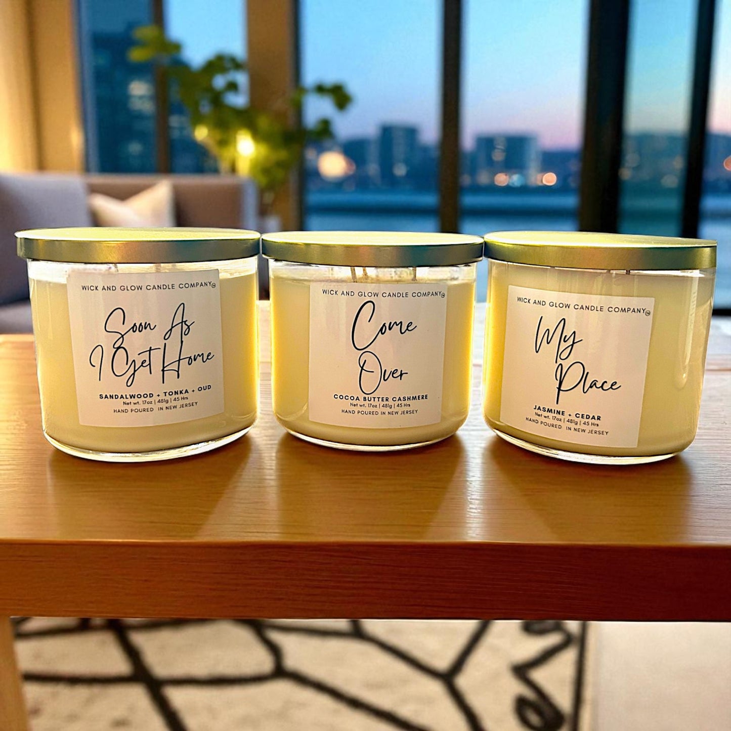 Three candles from Wick & Glow Candle Company on a wooden surface with a cityscape in the background.