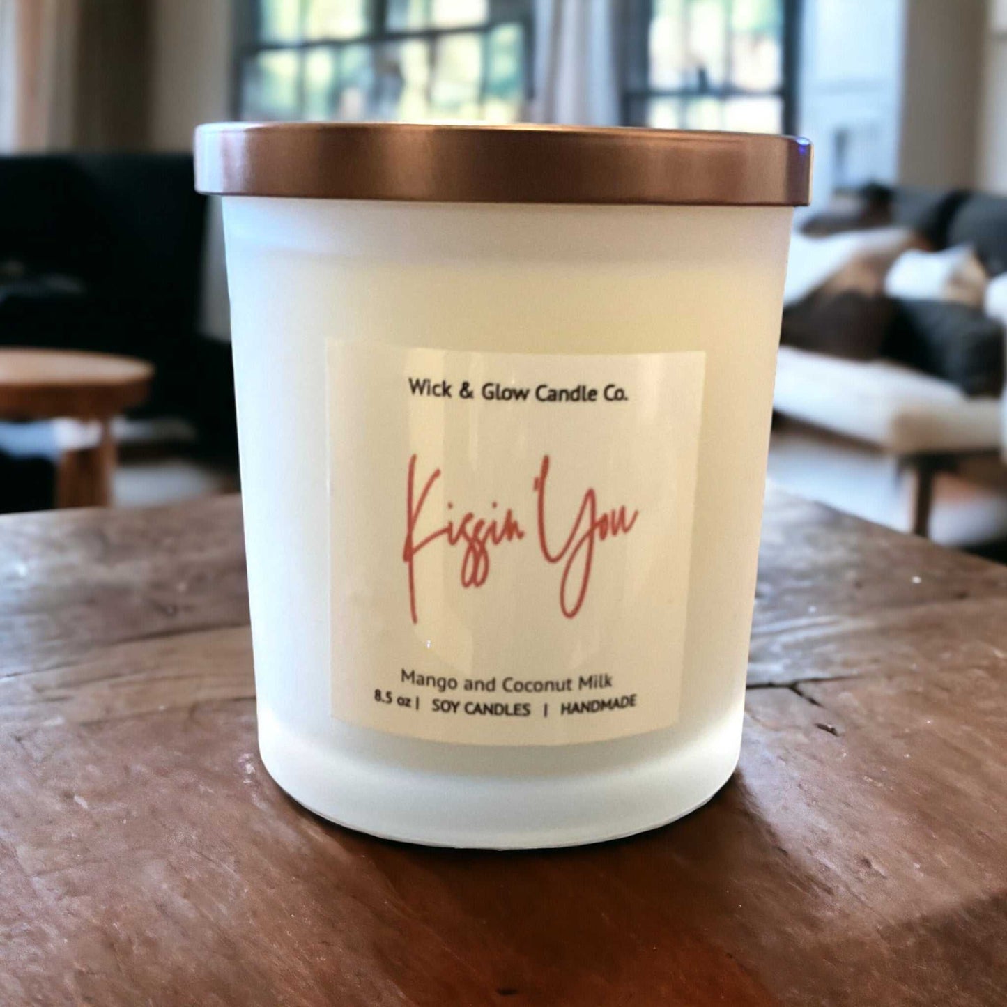 White candle with bronze lid on a table with a label that reads Kissin You in pink