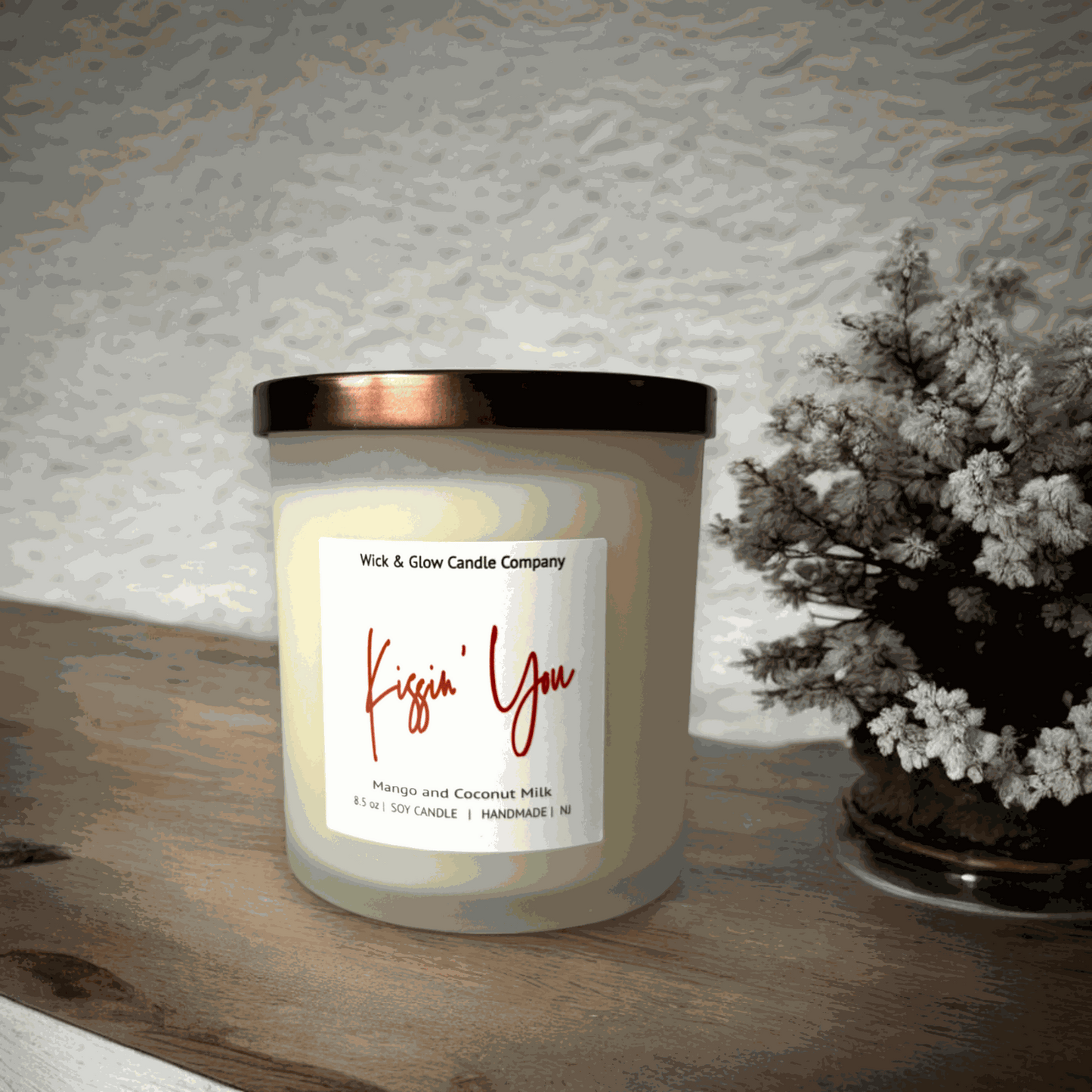 A white candle in a frosted glass jar with a bronze lid with a white label from Wick and Glow Candle Company on a wooden table with a plant in the background.