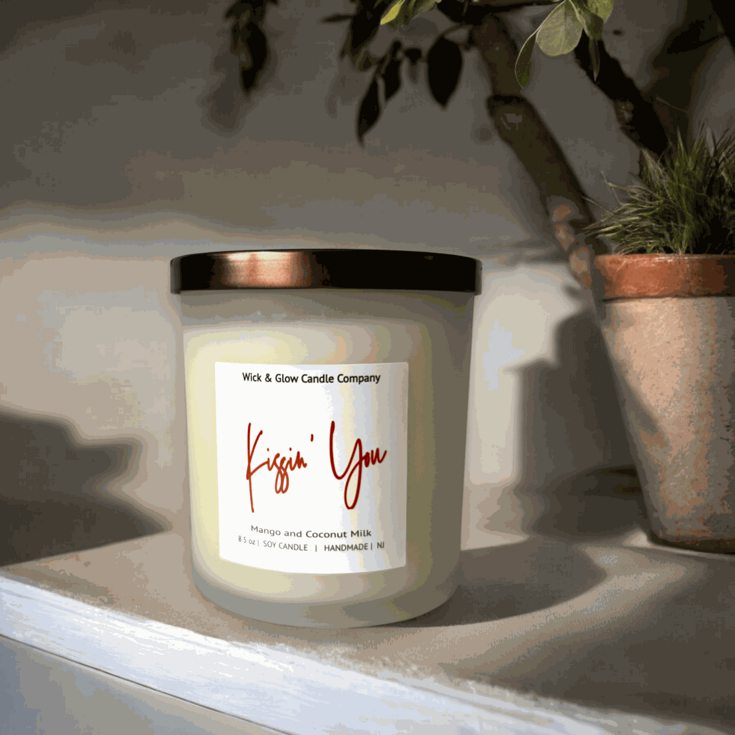 A white candle in a frosted glass jar with a bronze lid with a white label from Wick and Glow Candle Company on a white table with a plant in the background.