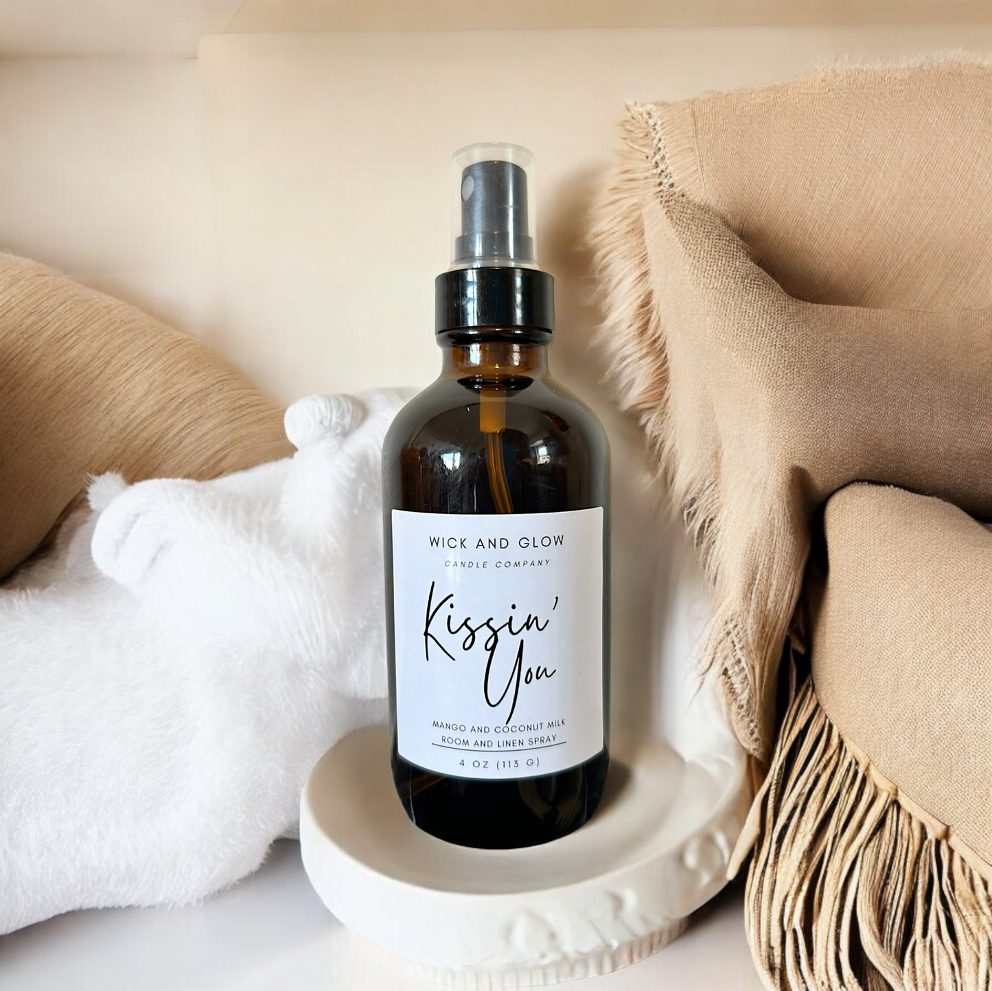 Amber spray bottle with a white label from the Wick and Glow Candle Company on a white table with white and tan blankets in the background ; the scent is mango and coconutmilk