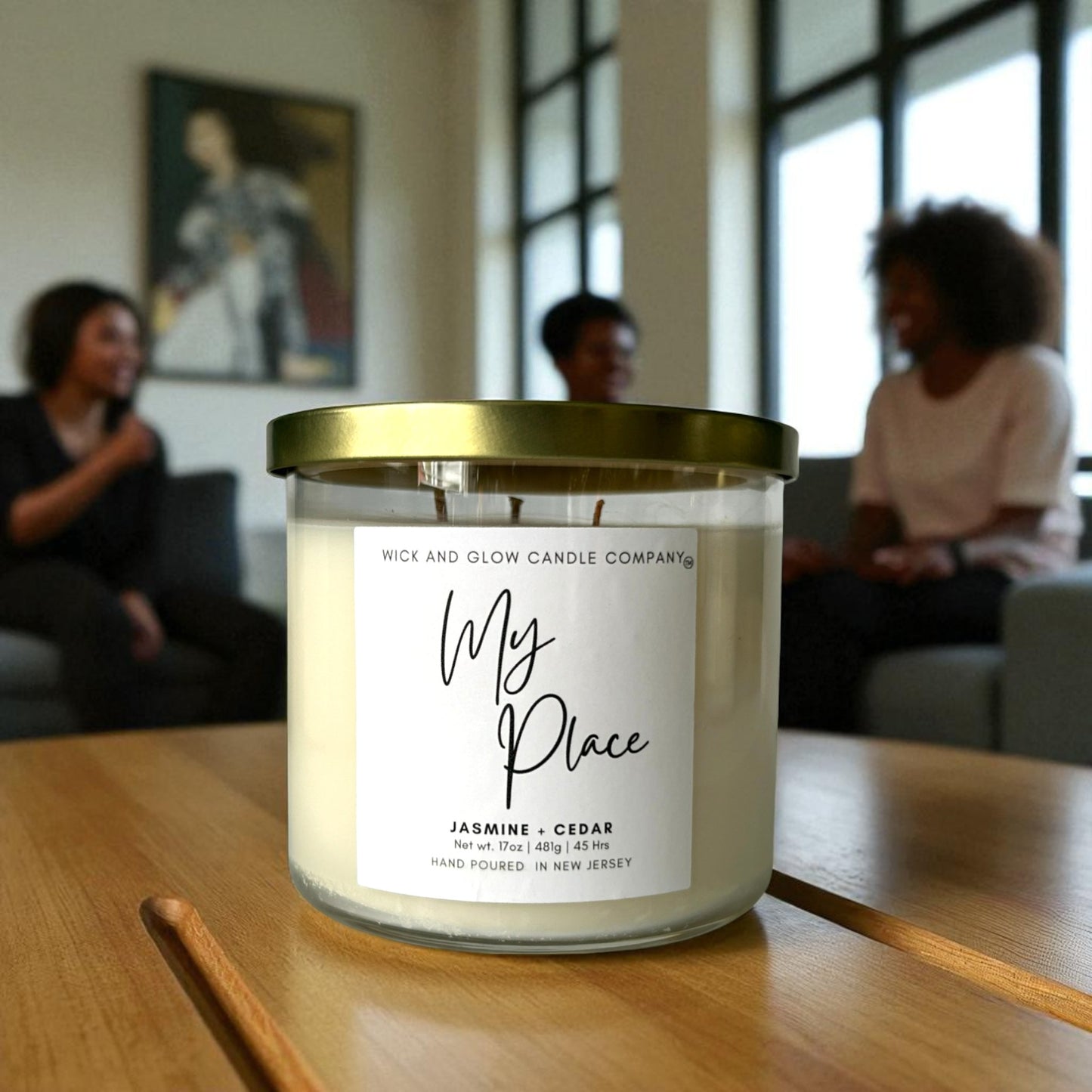 Candle labeled 'My Place' by Wick and Glow Candle Company on a wooden table with people in the background.