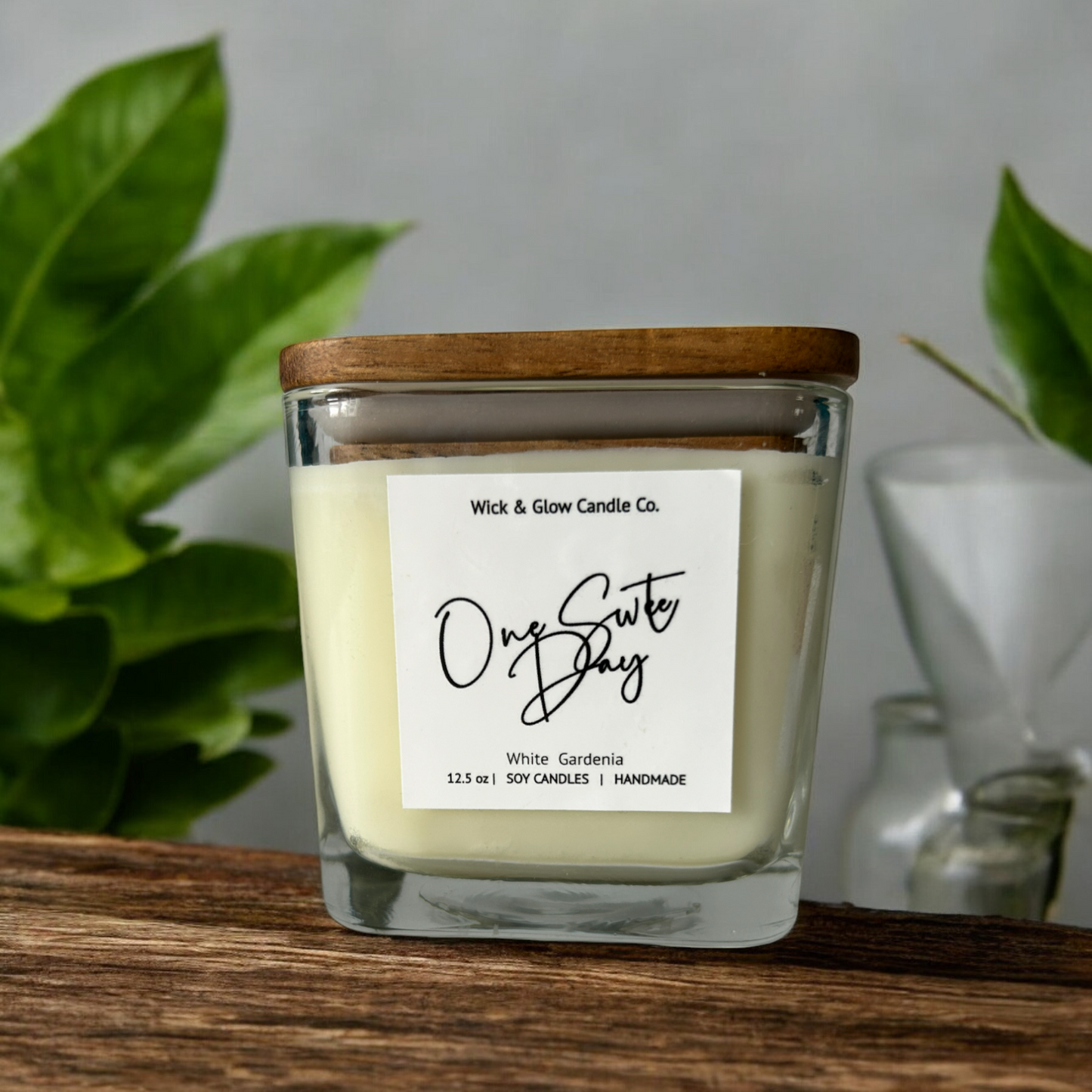 White candle in a clear square jar with a wooden lid on a wooden table with white background surrounded by plants.