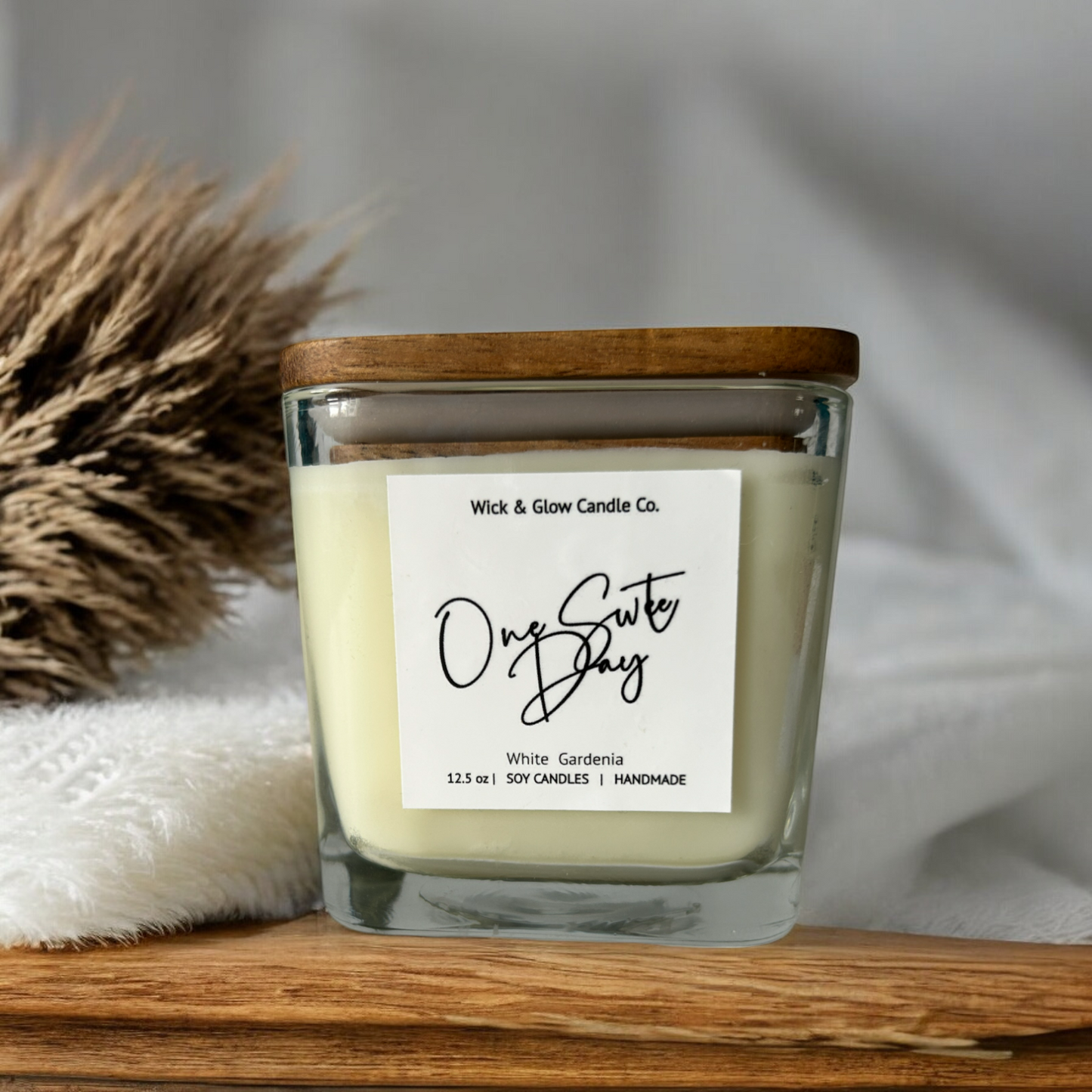 White candle in a clear square jar with a wooden lid on a wooden table with a white background and a brown plant.