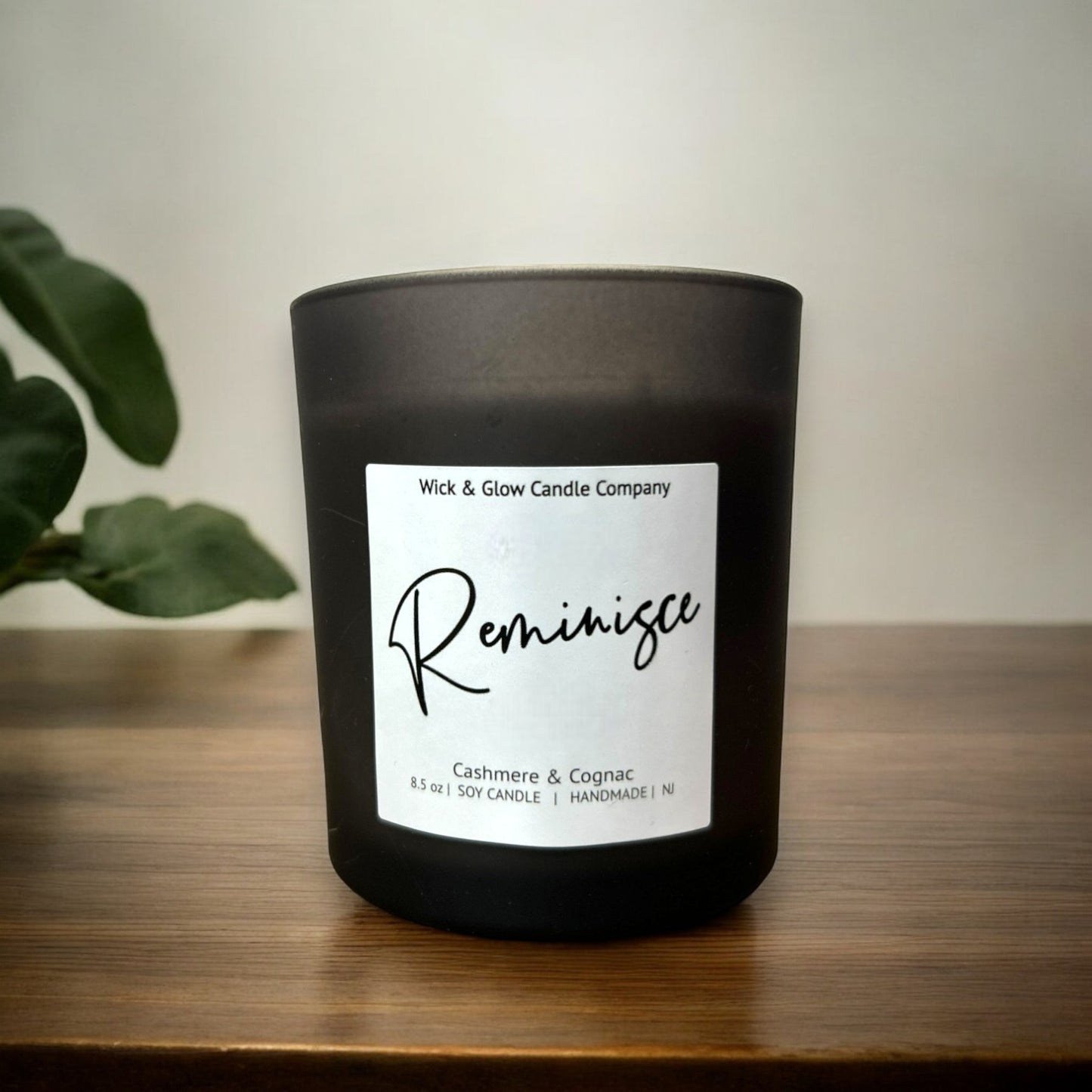 Black candle with a label on a wooden surface, featuring 'Wick & Glow Candle Company' and 'Reminisce'.