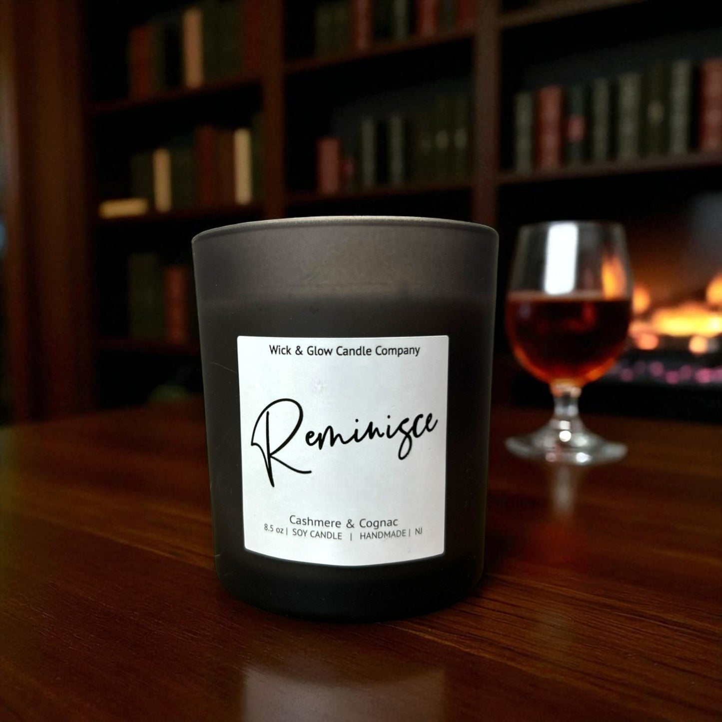Candle labeled 'Reminisce' from Wick & Glow Candle Company on a wooden surface with a glass of wine and bookshelf in the background.