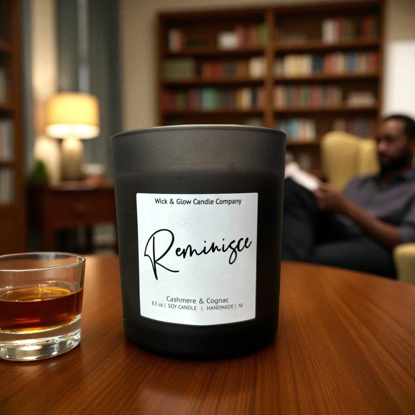 Black candle labeled 'Reminisce' on a wooden table with a blurred background of a person and bookshelves.