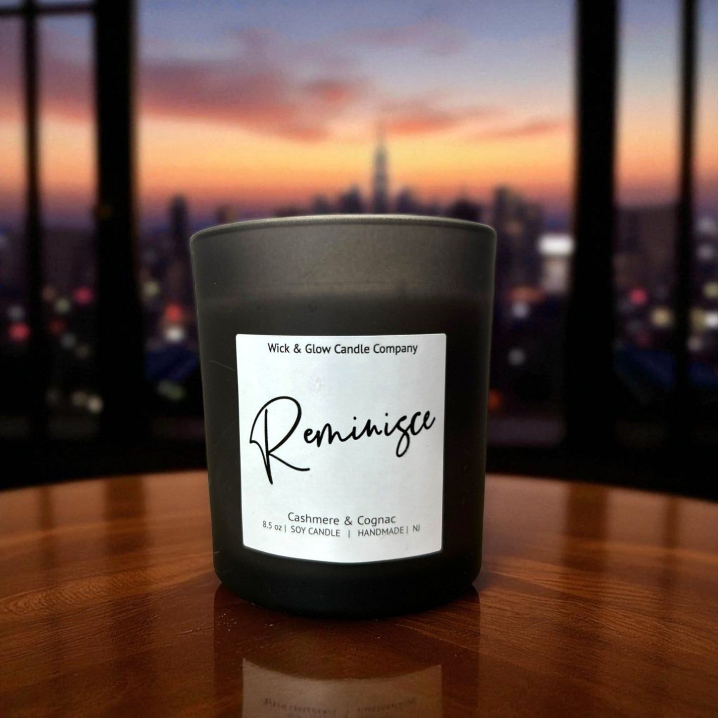 Black candle labeled 'Reminisce' on a wooden surface with a cityscape background during sunset.