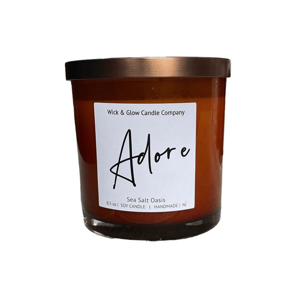 Candle in brown jar with white label that reads Adore on a white background