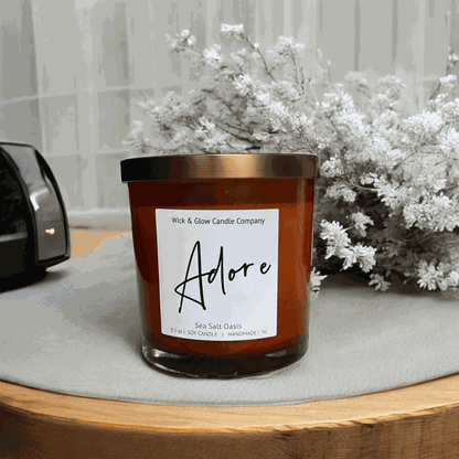 Wick and Glow soy wax candle in an amber glass jar with a  bronze lid on a wooden table. Scent notes: Sea Salt, Cardamom, Palm, Light Musk, Amber, hints of lilac; lilac scented candle