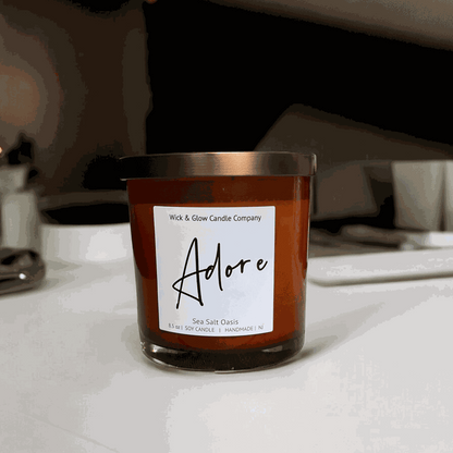 Wick and Glow soy wax candle in an amber glass jar with a  bronze lid on a white counter.  Scent notes: Sea Salt, Cardamom, Palm, Light Musk, Amber, hints of lilac; lilac scented candle