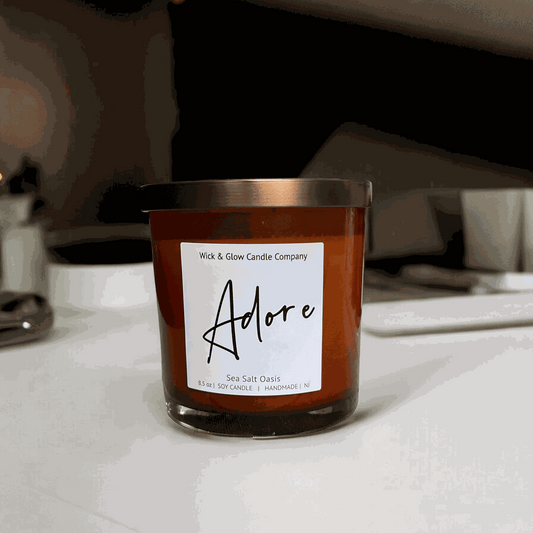 Wick and Glow soy wax candle in an amber glass jar with a  bronze lid on a white counter.  Scent notes: Sea Salt, Cardamom, Palm, Light Musk, Amber, hints of lilac; lilac scented candle