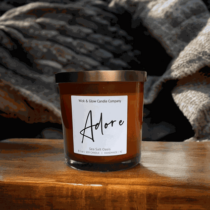 Wick and Glow soy wax candle in an amber glass jar with a  bronze lid on a wooden table. Scent notes: Sea Salt, Cardamom, Palm, Light Musk, Amber, hints of lilac; lilac scented candle