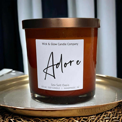 Wick and Glow soy wax candle in an amber glass jar with a  bronze lid on a metal tray. Scent notes: Sea Salt, Cardamom, Palm, Light Musk, Amber, hints of lilac; lilac scented candle