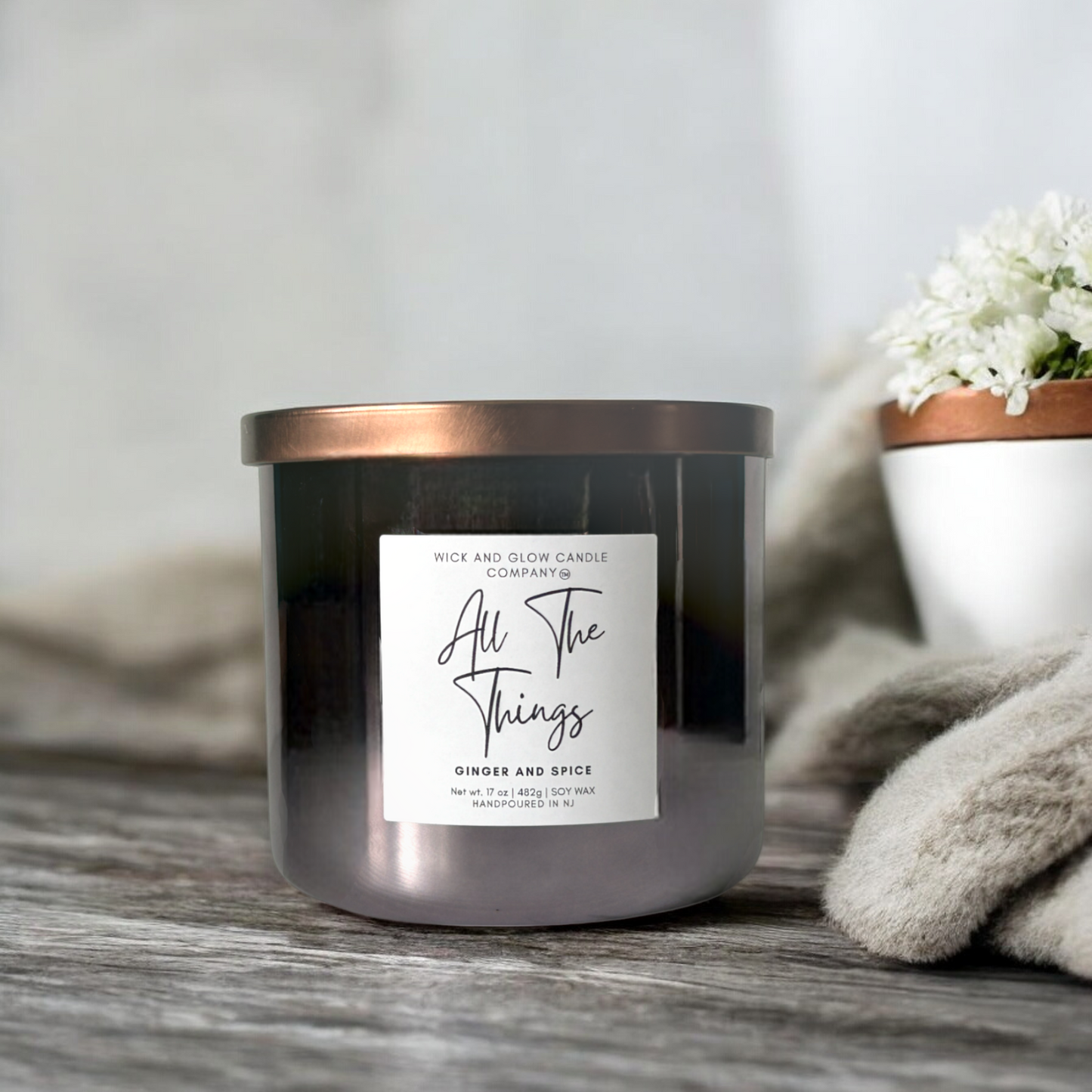 Wick and Glow luxury soy wax candle in a gray glass container with a bronze lid on a gray wooden table  with a plant in the background. The scent is Ginger and spice