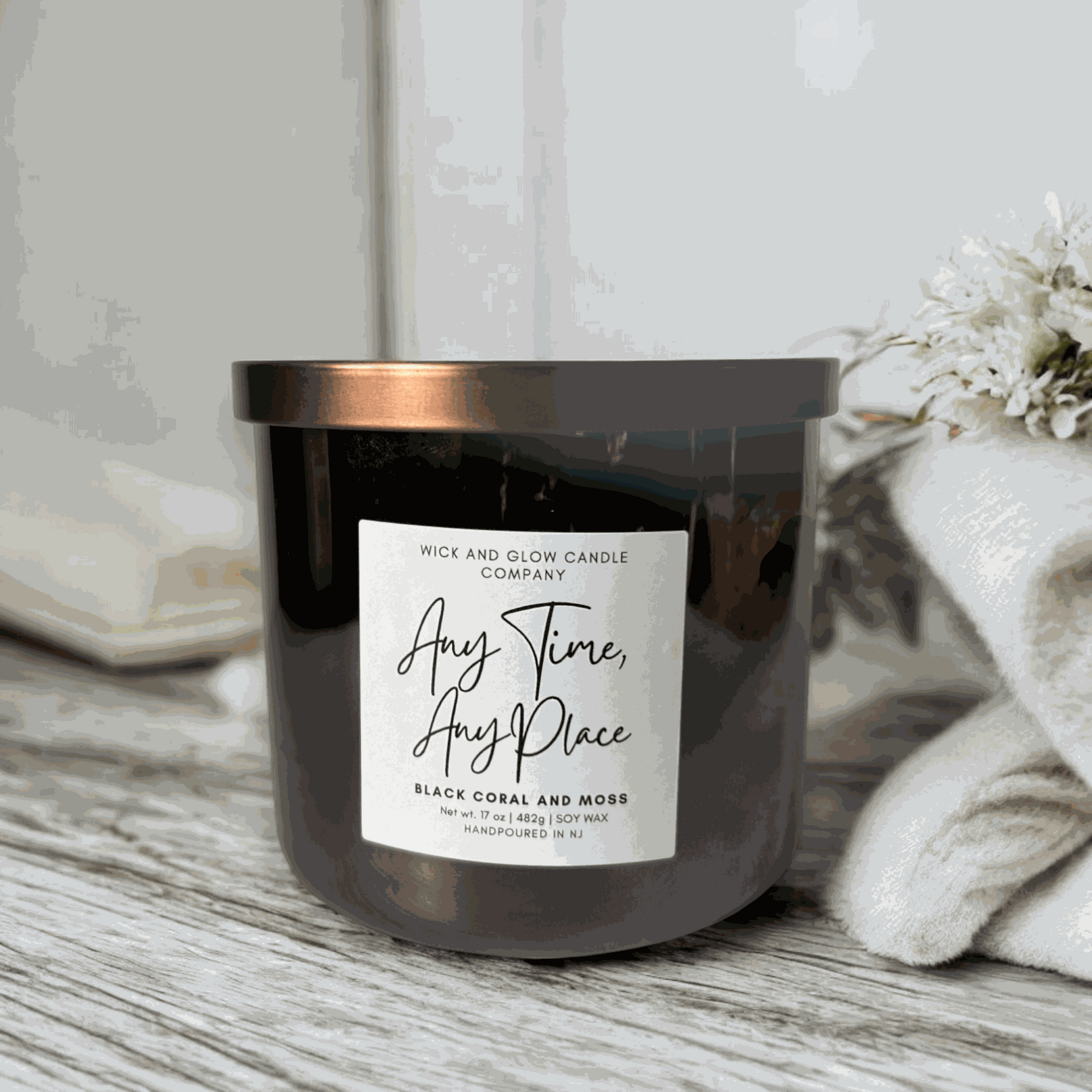 Wick and Glow's large luxury soy wax candle in a gray glass container with a bronze lid on a light grey  table with a blanket