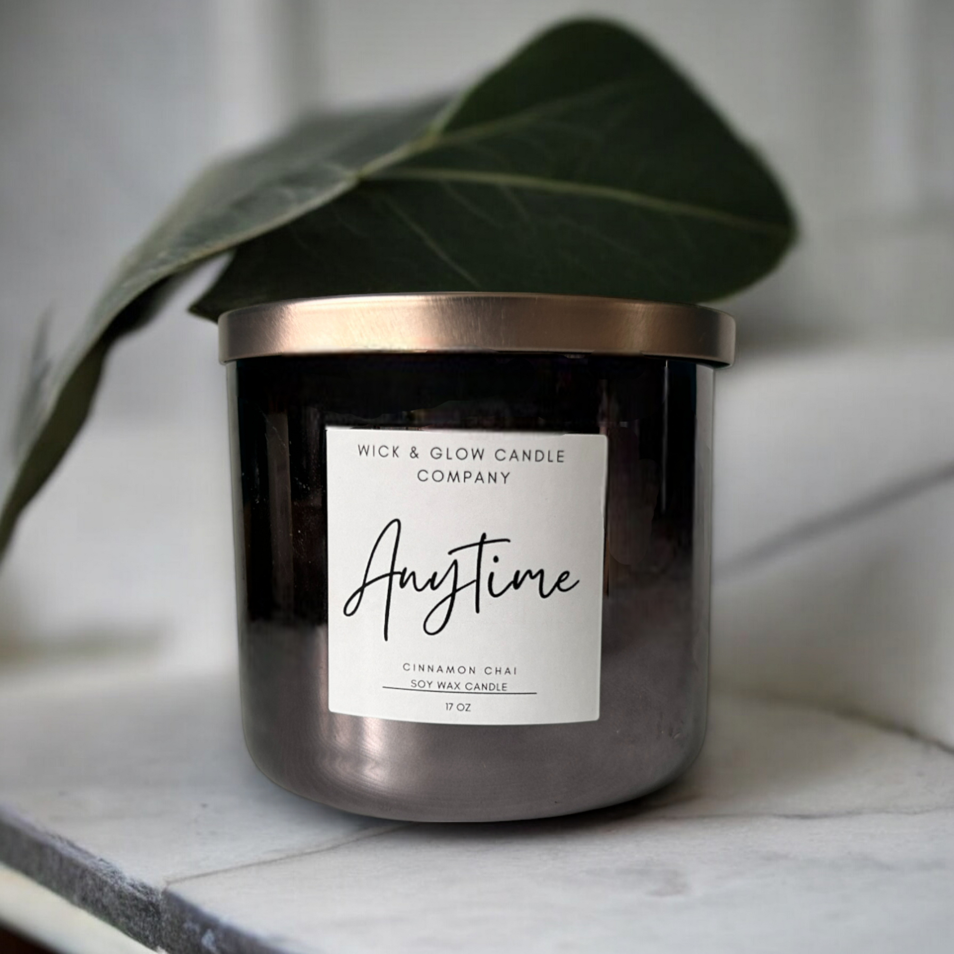 Wick and Glow's luxury soy wax candle in a gray jar with a bronze lid on a marble table with a large leaf in the background