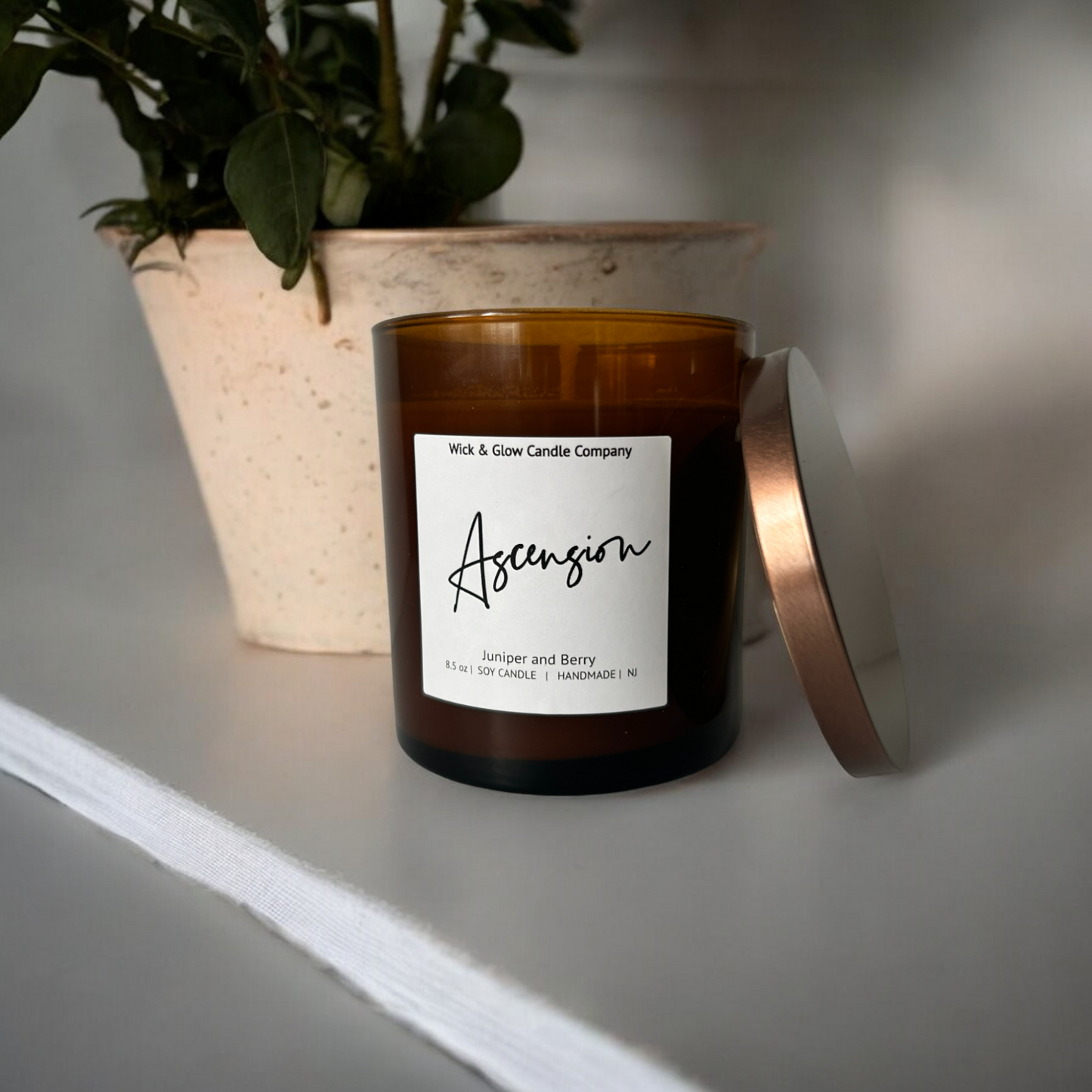 Wick and Glow luxury soy wax candle in an amber glass container on a white table with a plant behind it. The scent is Juniper and Berry.