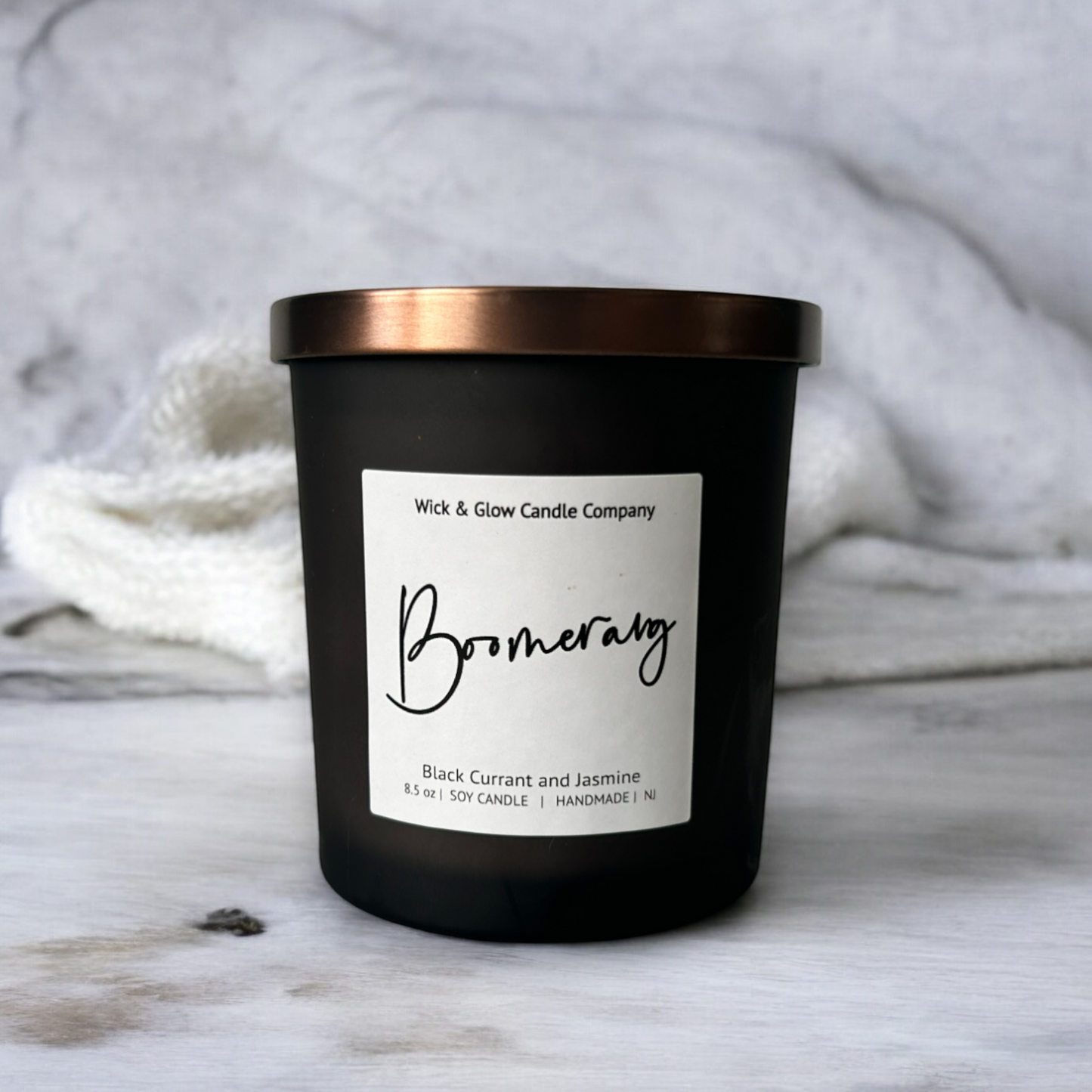 Wick and Glow luxury soy wax candle in a black glass container with a bronze lid and white label on a white marble table with a white background