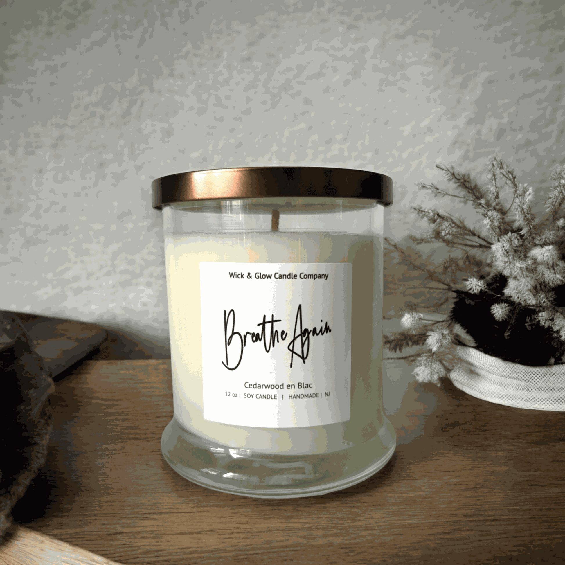 Wick and Glow's luxury soy wax candle in a clear glass container on a wooden table with wooden table with white flowers. The scent is Cedarwood.