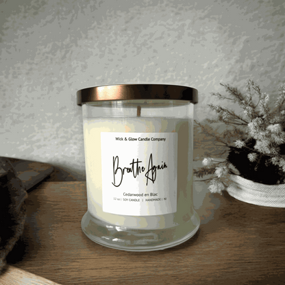 Wick and Glow's luxury soy wax candle in a clear glass container on a wooden table with wooden table with white flowers. The scent is Cedarwood.