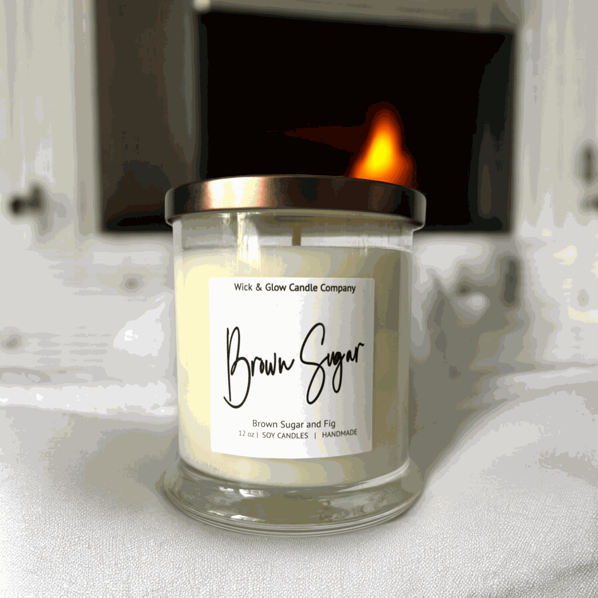 Wick and Glow's luxury soy wax candle in a clear glass container  on a white sheet with a fireplace in the background. The scent is Brown Sugar and Fig.