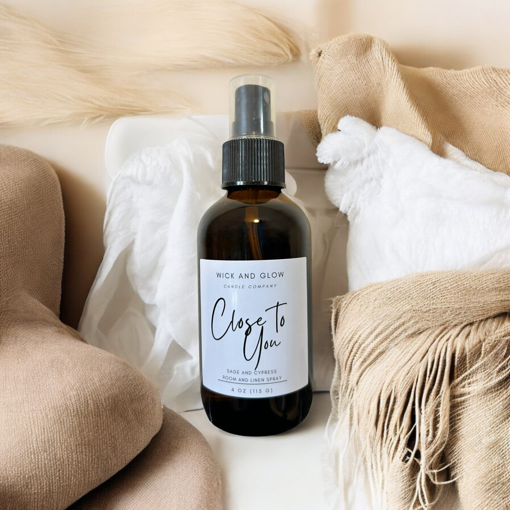 Amber spray bottle  with a white label from the Wick and Glow Candle Company on a white table with tan blankets in the background ; the scent is sage and cypress