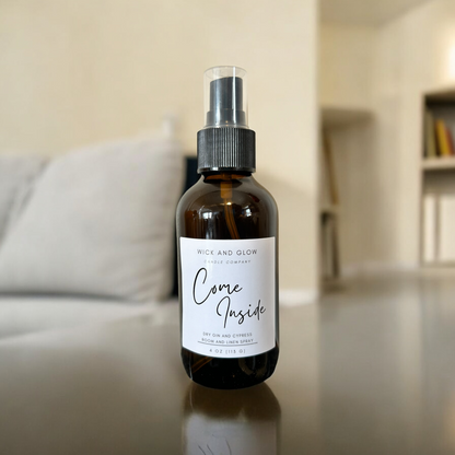 Amber spray bottle  with a white label from the Wick and Glow Candle Company on a glass table with a white couch; the scent is dry gin and cypress