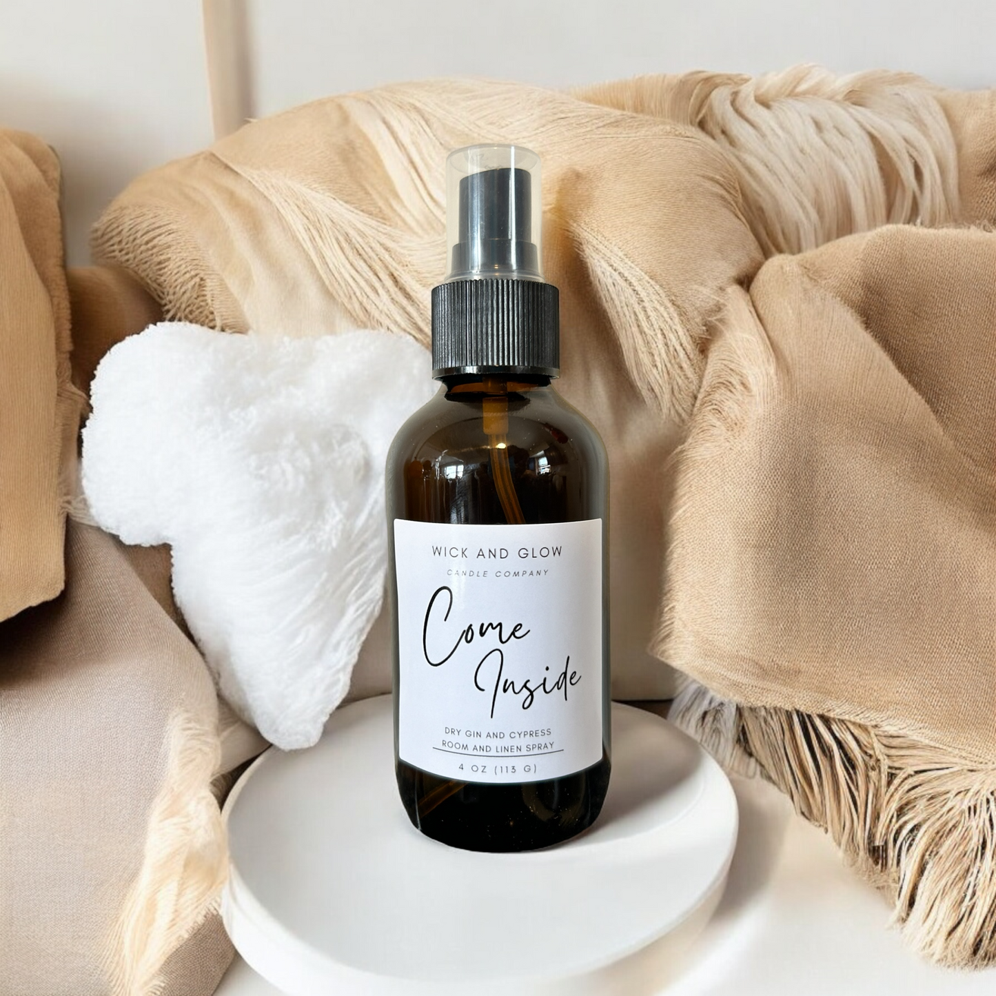 Amber spray bottle  with a white label from the Wick and Glow Candle Company on a white pedestal with tan blankets in the background ; the scent is dry gin and cypress