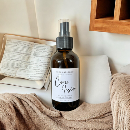 Amber spray bottle  with a white label from the Wick and Glow Candle Company on a white pedestal with tan blankets in the background ; the scent is dry gin and cypress