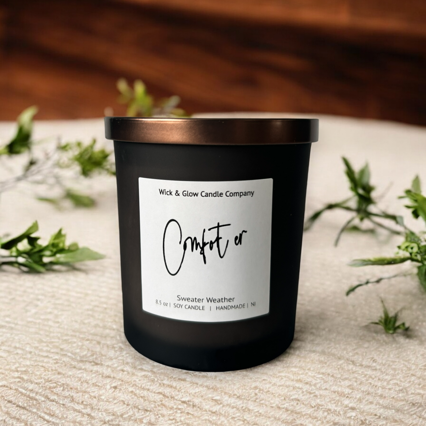 Wick and Glow's luxury soy wax candle in a frosted black glass container with a bronze lid on a tan table cloth with plants in the background.