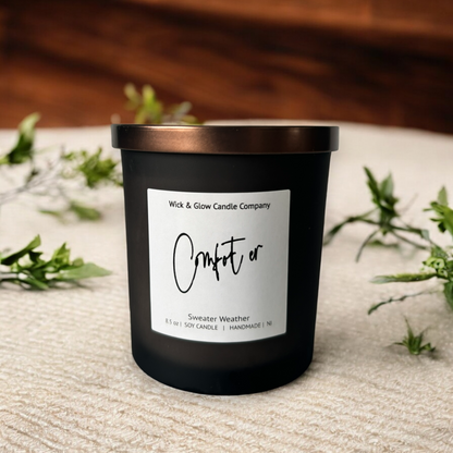 Wick and Glow's luxury soy wax candle in a frosted black glass container with a bronze lid on a tan table cloth with plants in the background.