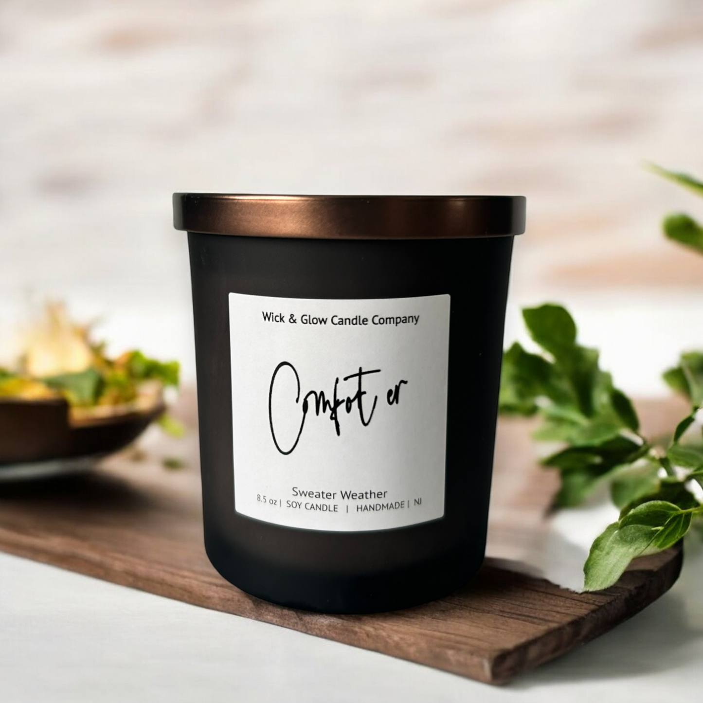 Wick and Glow's luxury soy wax candle in a frosted black glass container with a bronze lid on a wood  tray with plants in the background.