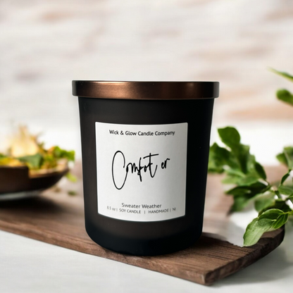 Wick and Glow's luxury soy wax candle in a frosted black glass container with a bronze lid on a wood  tray with plants in the background.
