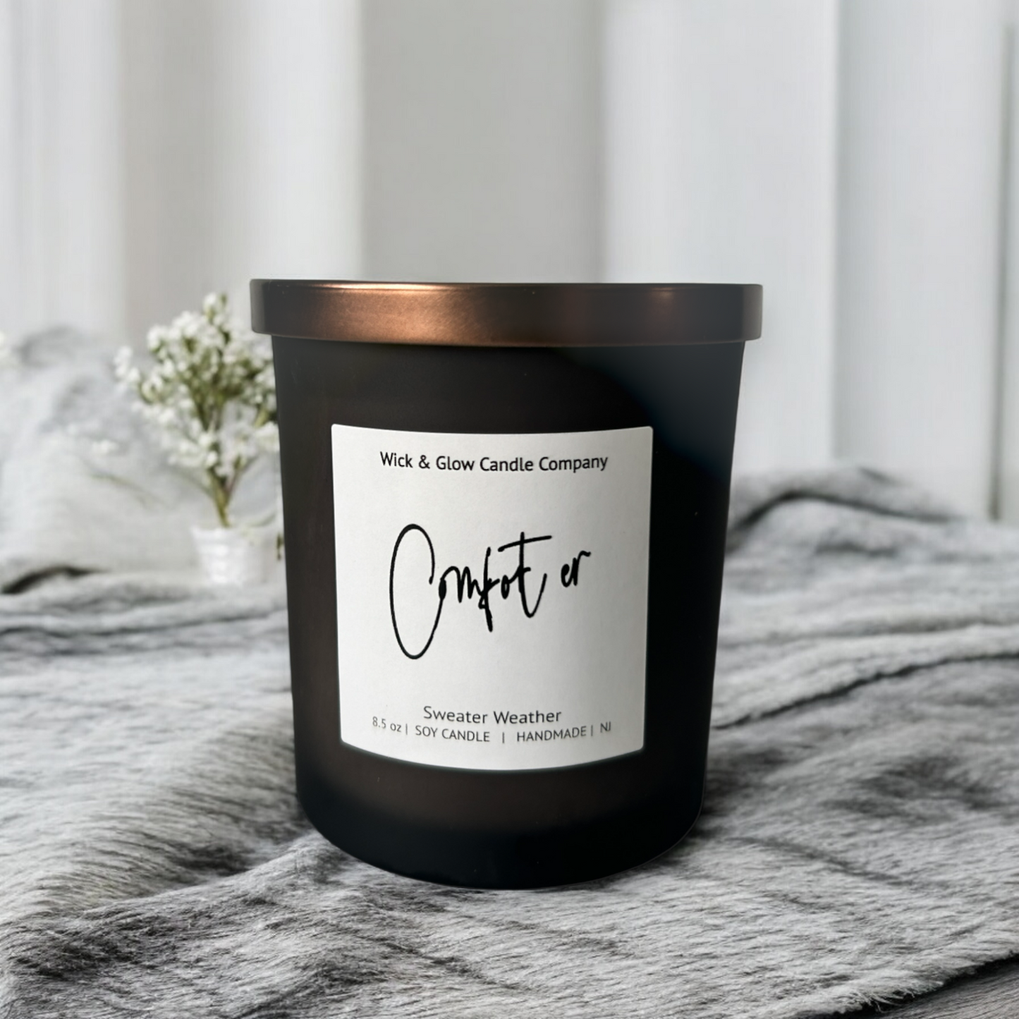 Wick and Glow's luxury soy wax candle in a frosted black glass container with a bronze lid on a gray blanket with plants in the background.