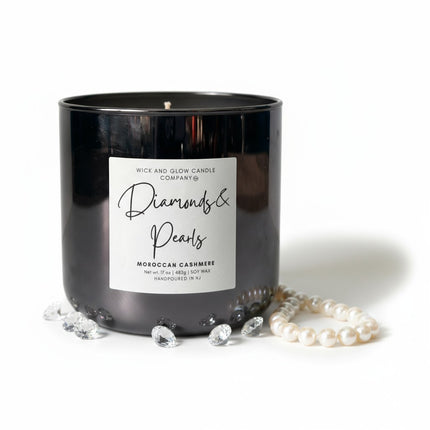 A black glass jar candle with a white label reading 'Diamonds & Pearls' by Wick and Glow Candle Company, styled with faux pearls and clear gemstone props on a white background.