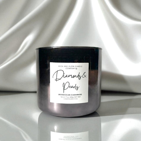 A dark metallic glass jar candle with a white label reading 'Diamonds & Pearls' and 'Rick and Glow Candle Company,' set against draped white satin fabric.
