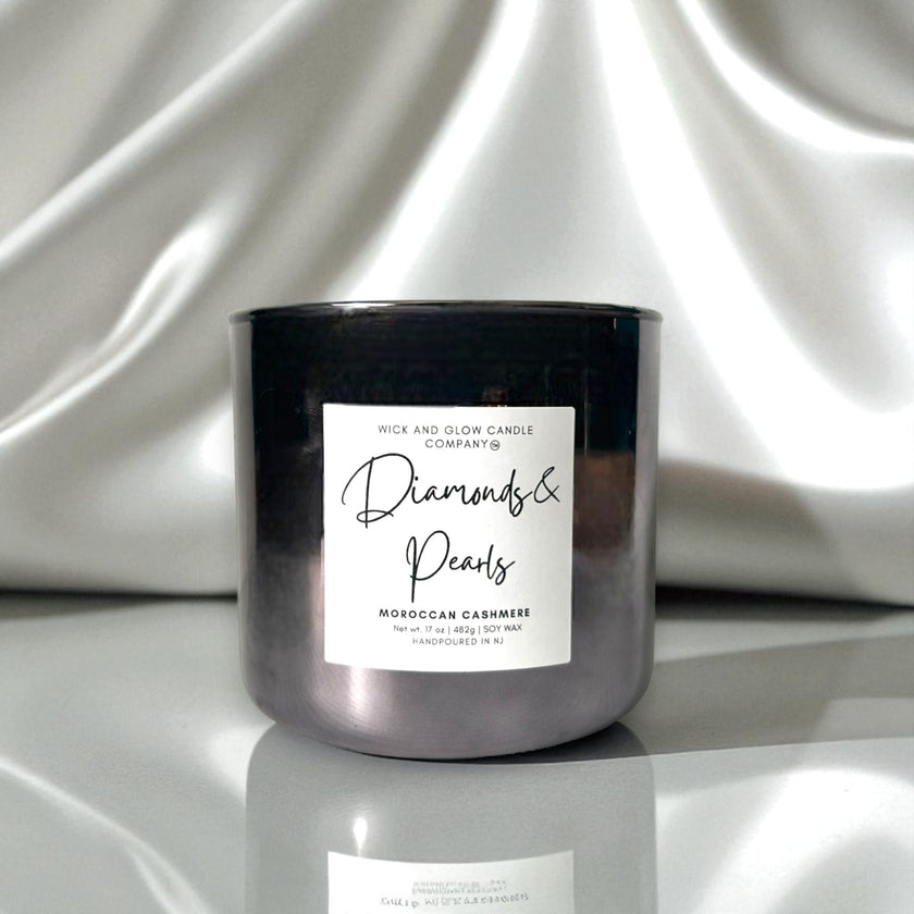 A dark metallic glass jar candle with a white label reading 'Diamonds & Pearls' and 'Rick and Glow Candle Company,' set against draped white satin fabric.