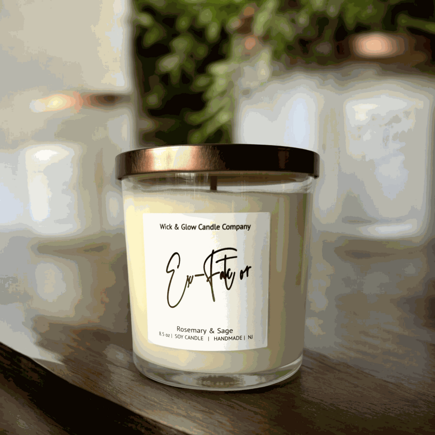 Rosemary and sage soy wax candle in a clear glass jar with a bronze lid on a wooden table with plants in the background.