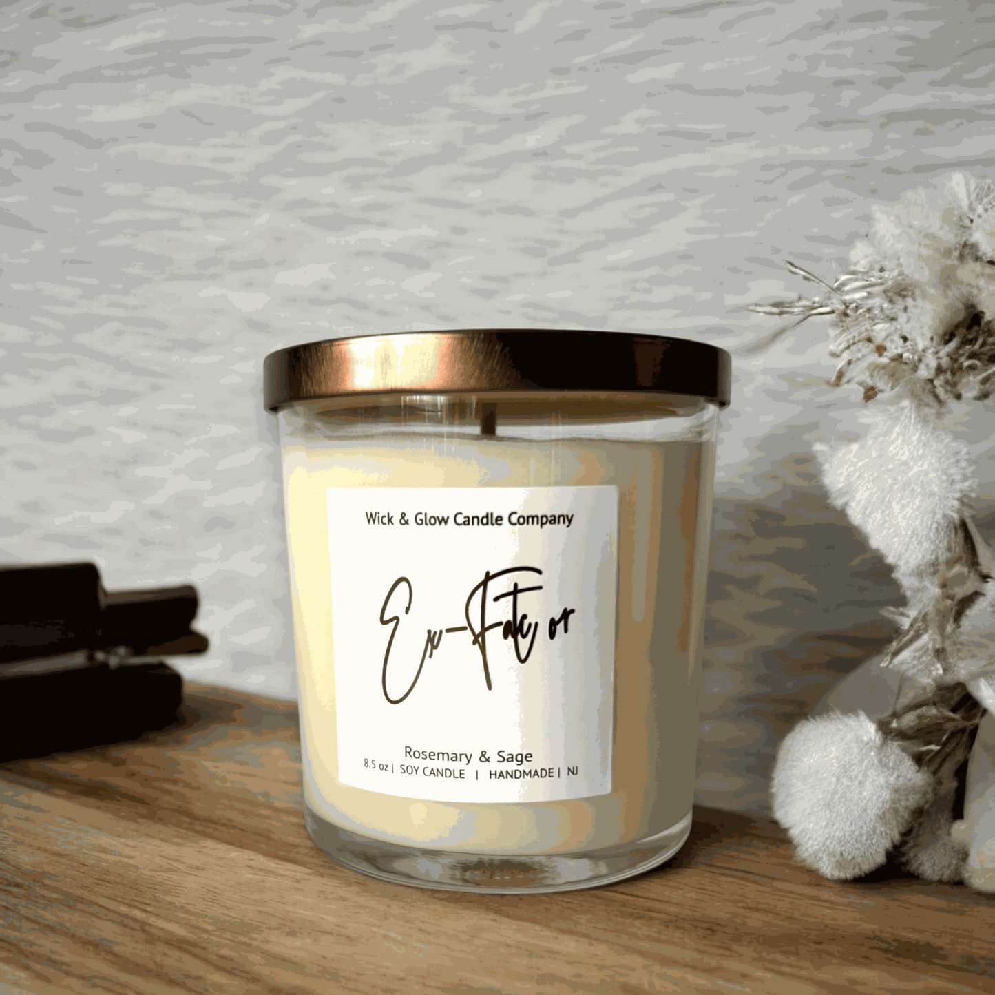 Rosemary and sage soy wax candle in a clear glass jar with a bronze lid on a rustic wooden table with a white background and white flowers from Wick and Glow Candle Company