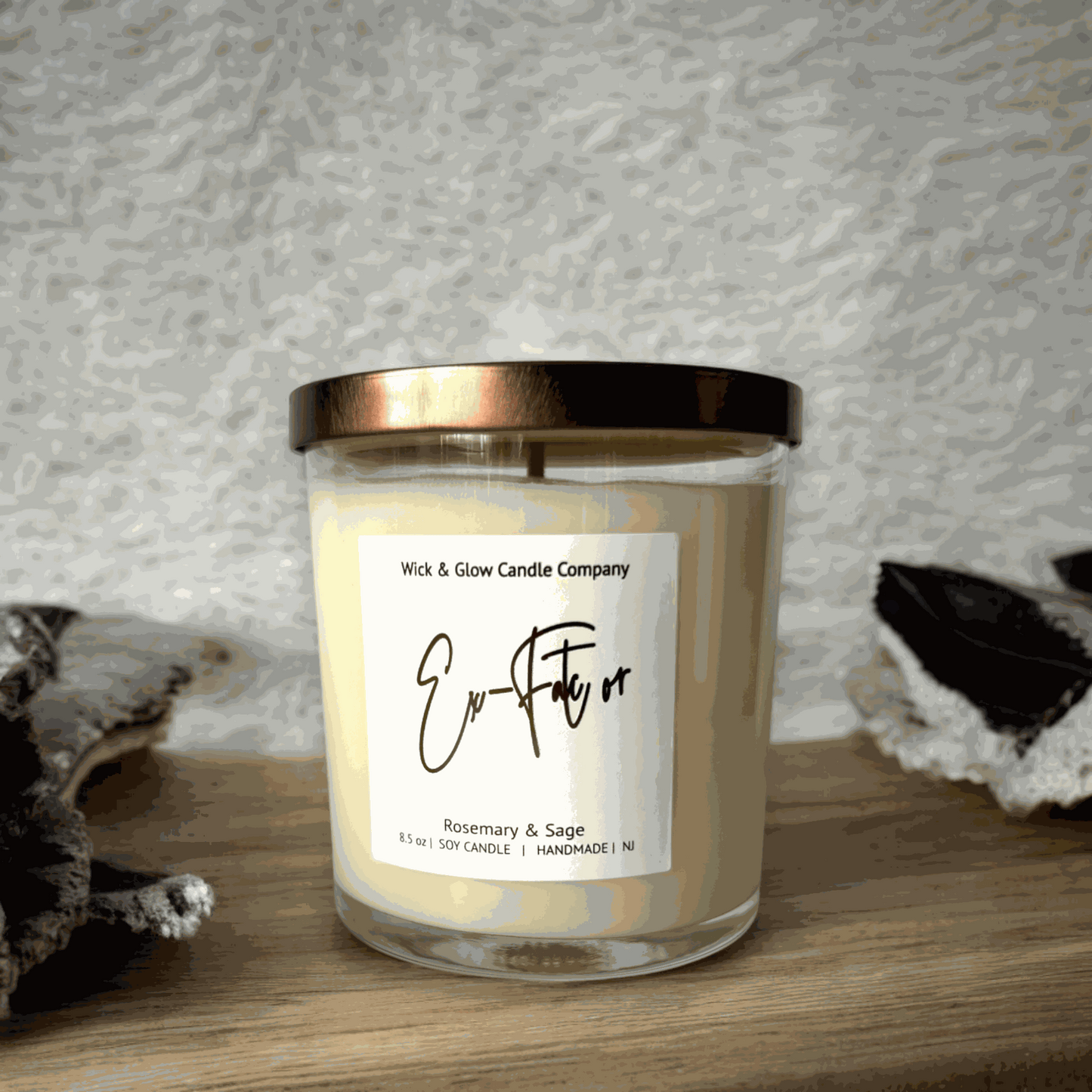 Rosemary and sage soy wax candle in a clear glass jar with a bronze lid on a rustic wooden table with a white background and white flowers from Wick and Glow Candle Company