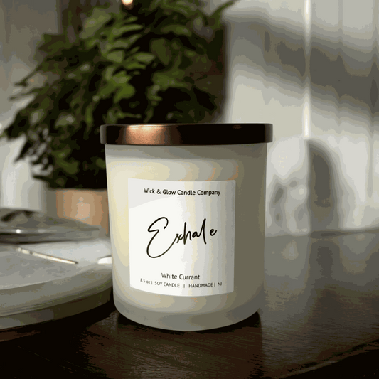 White currant soy wax candle in a frosted white glass jar with a bronze lid on a wooden table with with plants in the background from Wick and Glow Candle Company