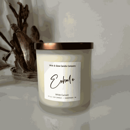 White currant soy wax candle from Wick and Glow Candle Company in a frosted white glass jar with a bronze lid on a white table with with a wooden sculpture in the background.