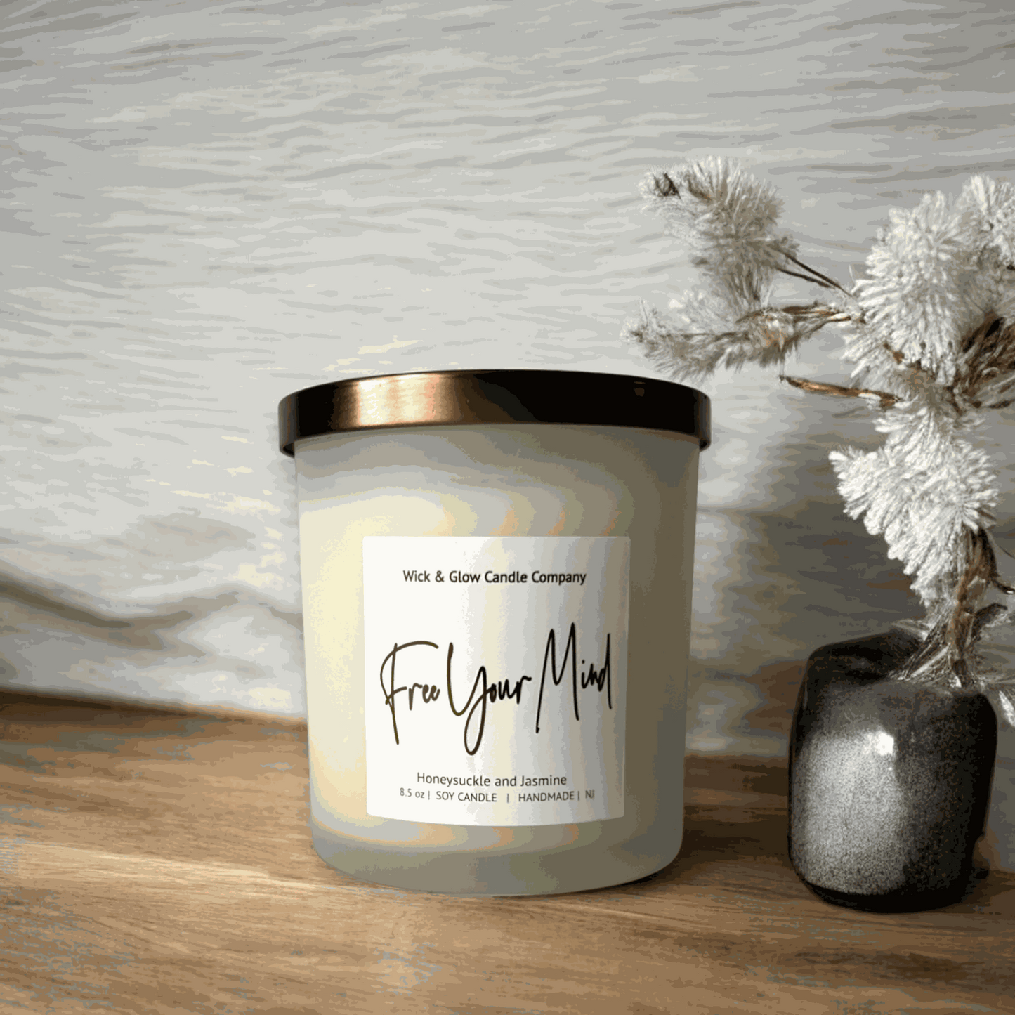 A white candle in a frosted glass jar with a bronze lid with a white label on a wooden table with a white background from Wick and Glow Candle Company.