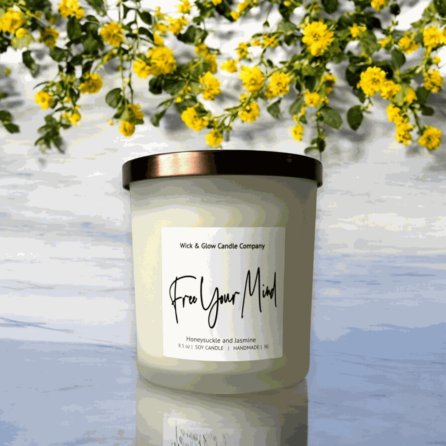 A white candle in a frosted glass jar with a bronze lid with a white label on a marble table with a white background and yellow flowers from Wick and Glow Candle Company.