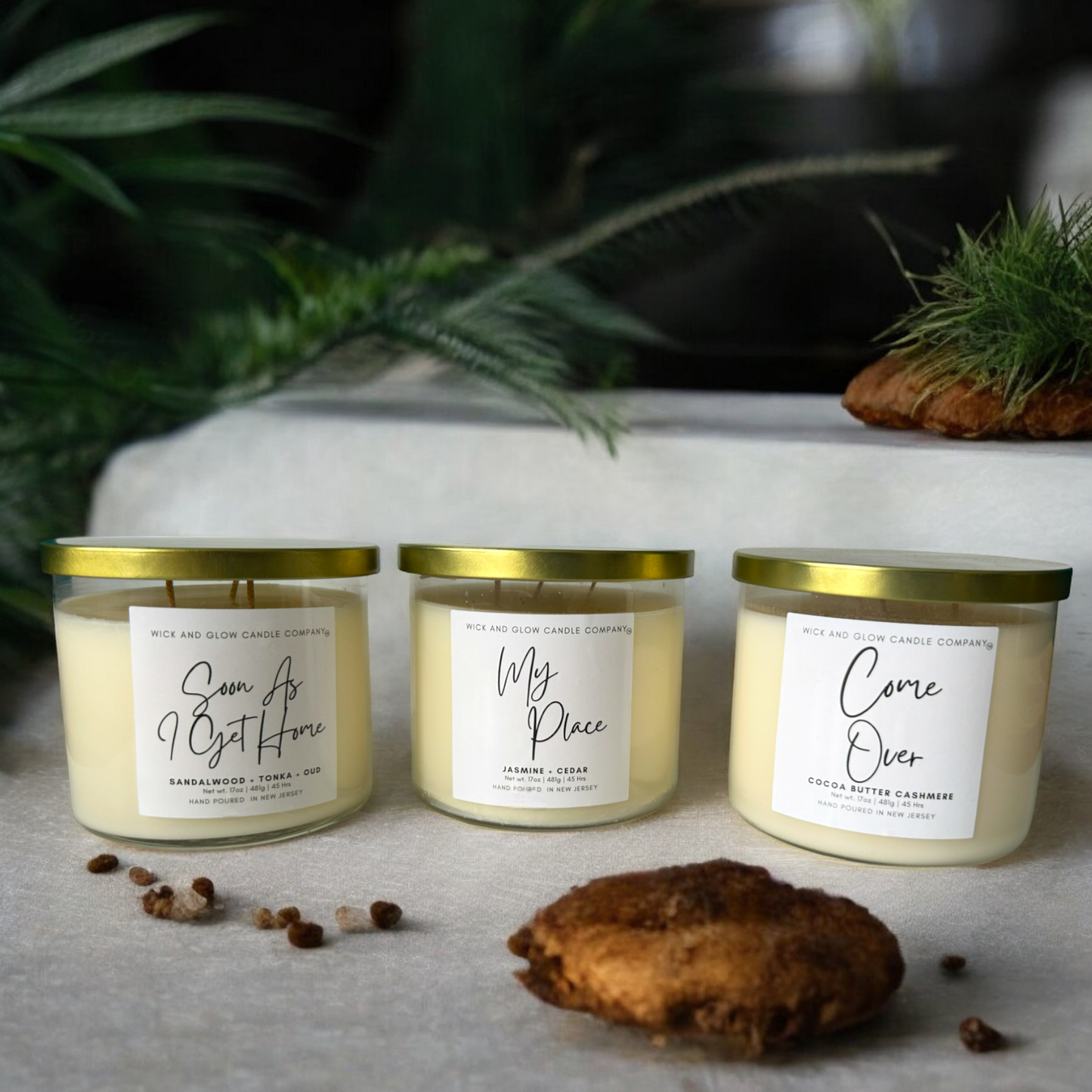 Three 17 oz luxury scented candles in clear jars with gold lids from Wick and Glow Candle Company on a white concrete table with branches and a wall in the background