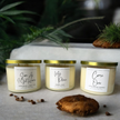Three candles with labels placed on a table with a cookie and greenery in the background, indicating a cozy, homey setting.