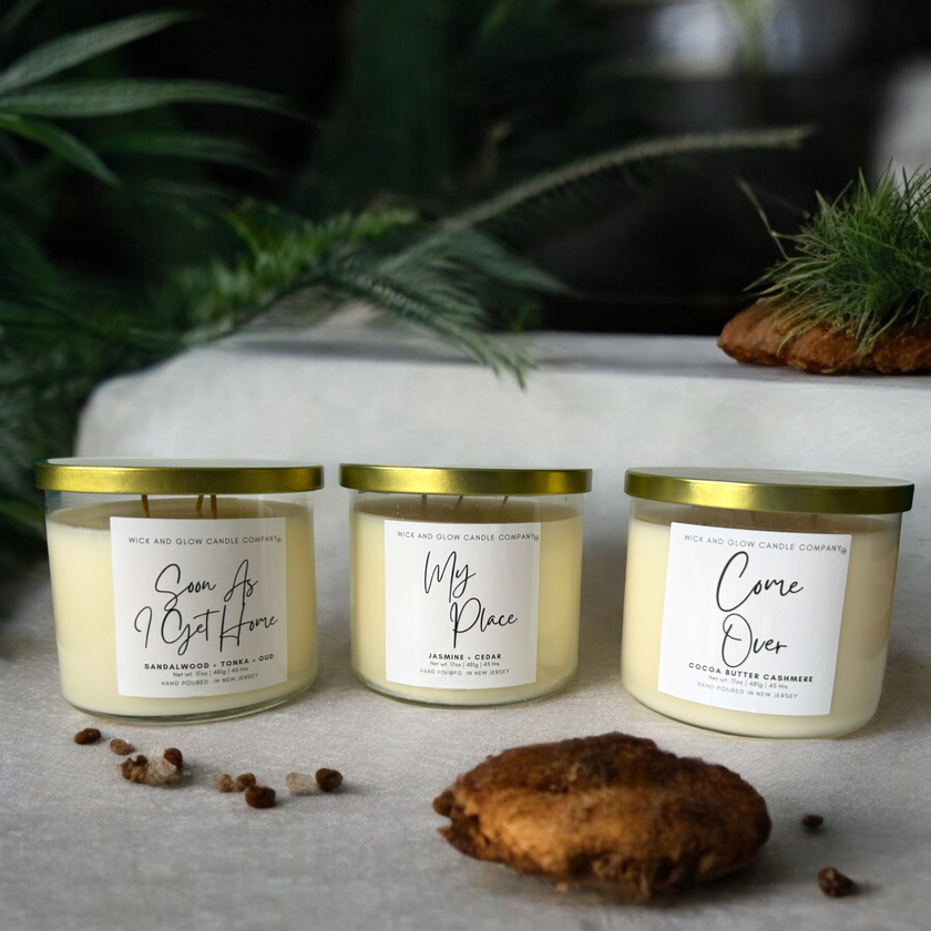 Three candles with labels placed on a table with a cookie and greenery in the background, indicating a cozy, homey setting.